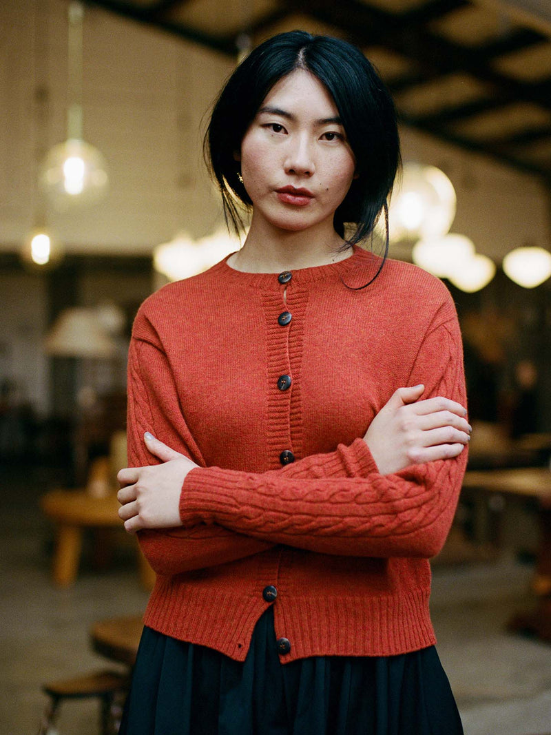 A petite woman with long dark hair stands indoors, arms crossed, wearing the fitted Kindred Cable Cardigan in Amber as she looks at the camera.