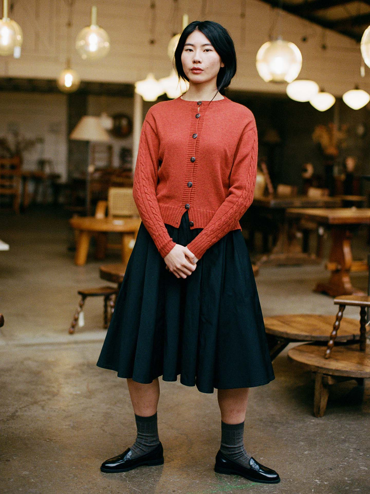 Petite woman indoors wearing the Kindred Cable Cardigan – Amber in a relaxed fit, paired with a black skirt, gray socks, and black shoes; hands clasped, with wooden furniture and hanging lights in the background.