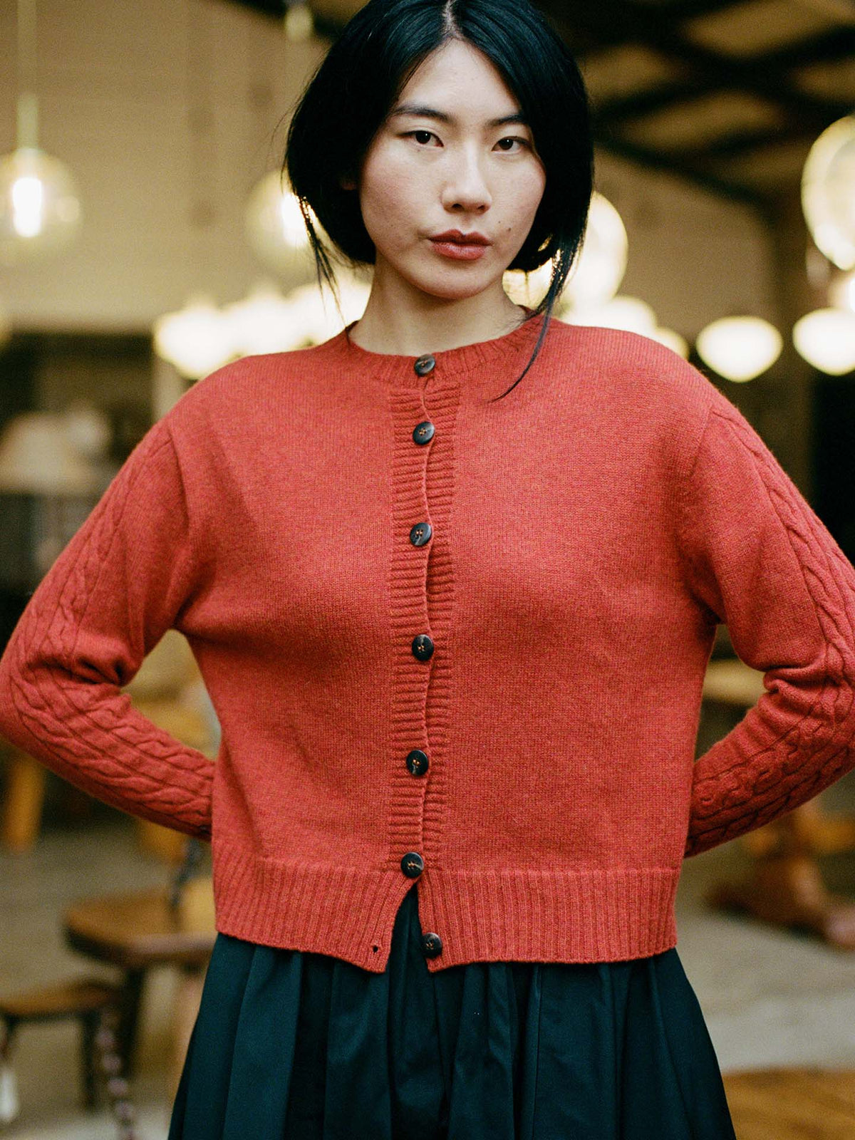 A petite woman with straight black hair wears the fitted Kindred Cable Cardigan in Amber and a black skirt, standing indoors with blurred lights and furniture in the background.