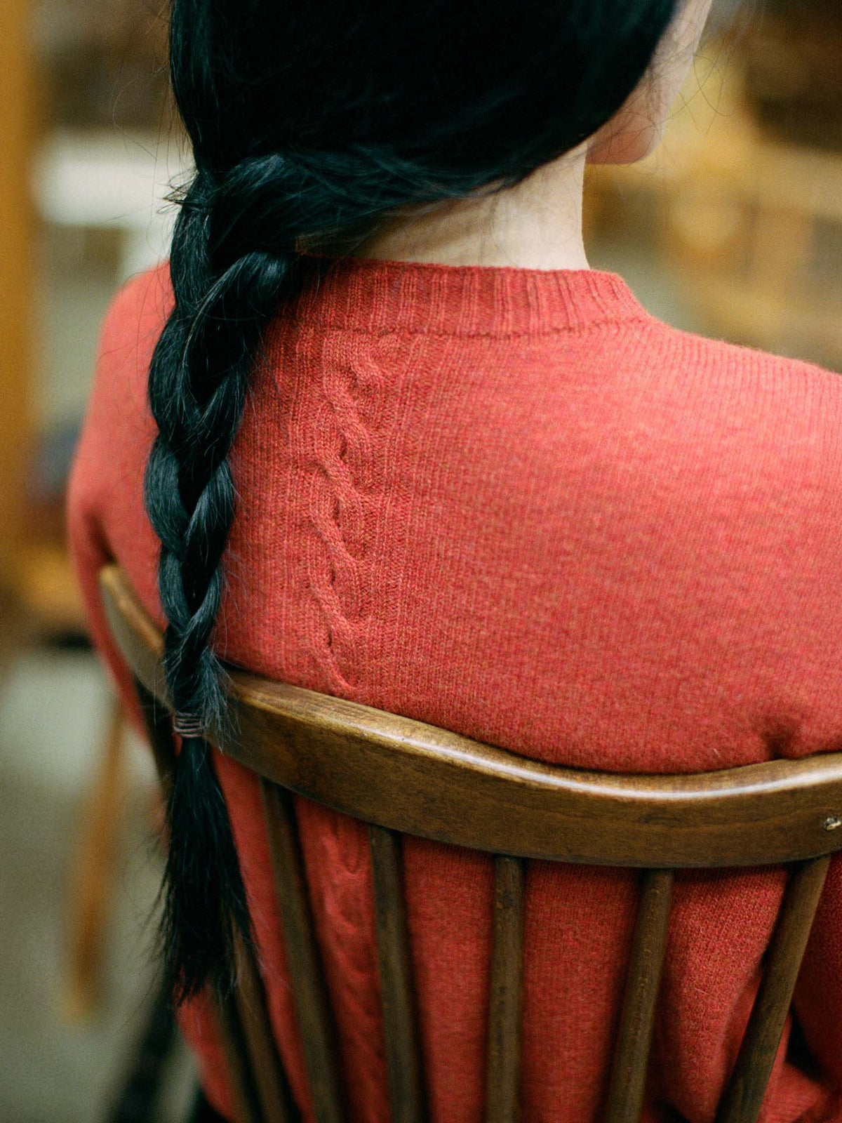 A petite person with long dark hair in a braid sits on a wooden chair, wearing the Kindred Cable Cardigan in Amber—a relaxed-fit red sweater featuring a cable knit pattern down the back.