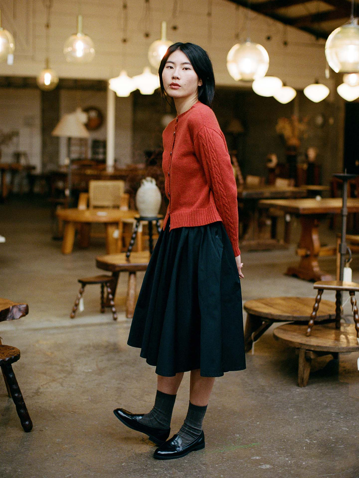 A petite woman stands indoors wearing the Kindred Cable Cardigan in Amber, paired with a black skirt, gray socks, and black shoes, surrounded by wooden tables and pendant lights.