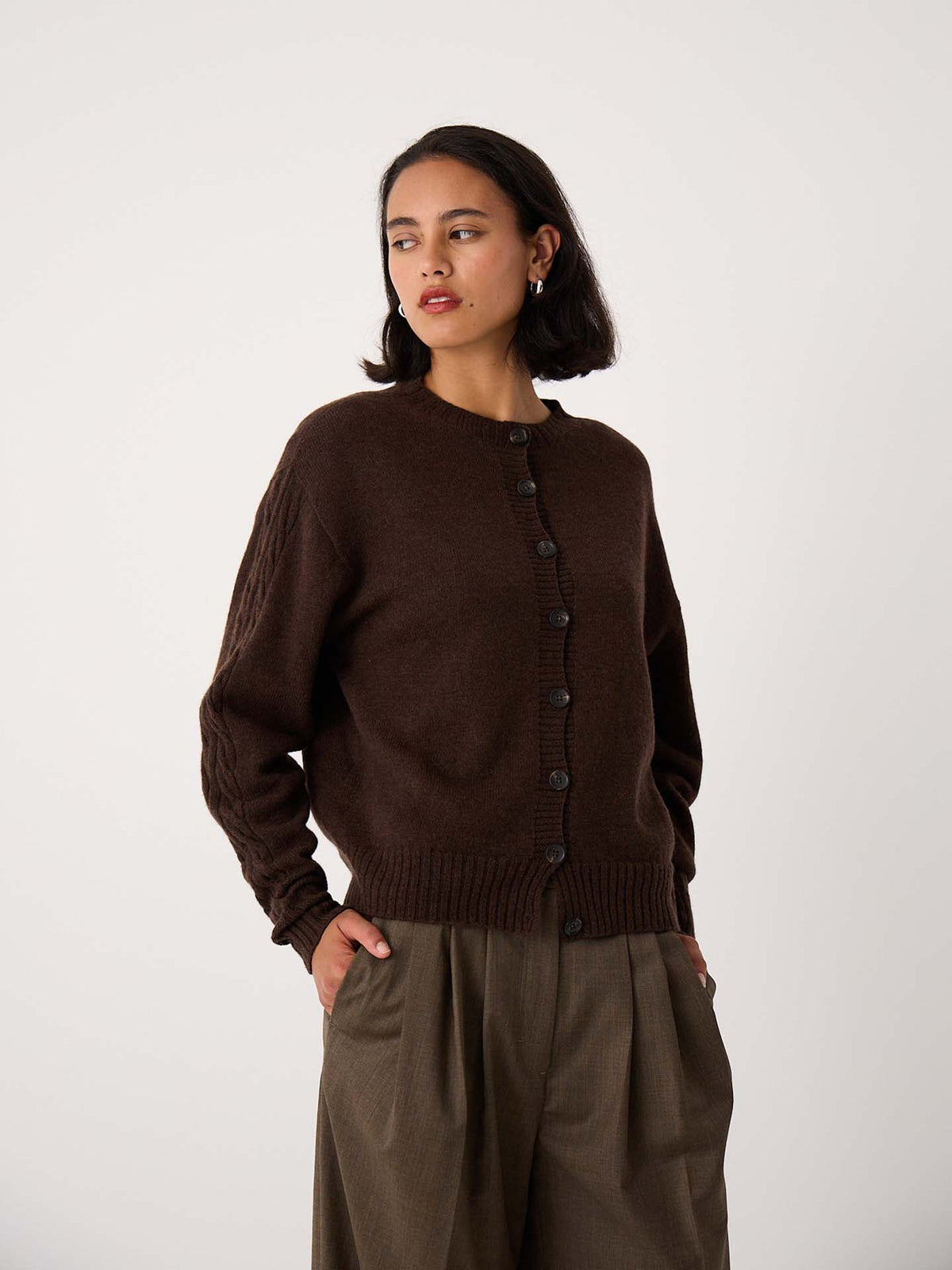 A woman wears the Kindred Cable Cardigan in Cocoa, styled with loose brown trousers, standing with one hand in her pocket against a plain white background.