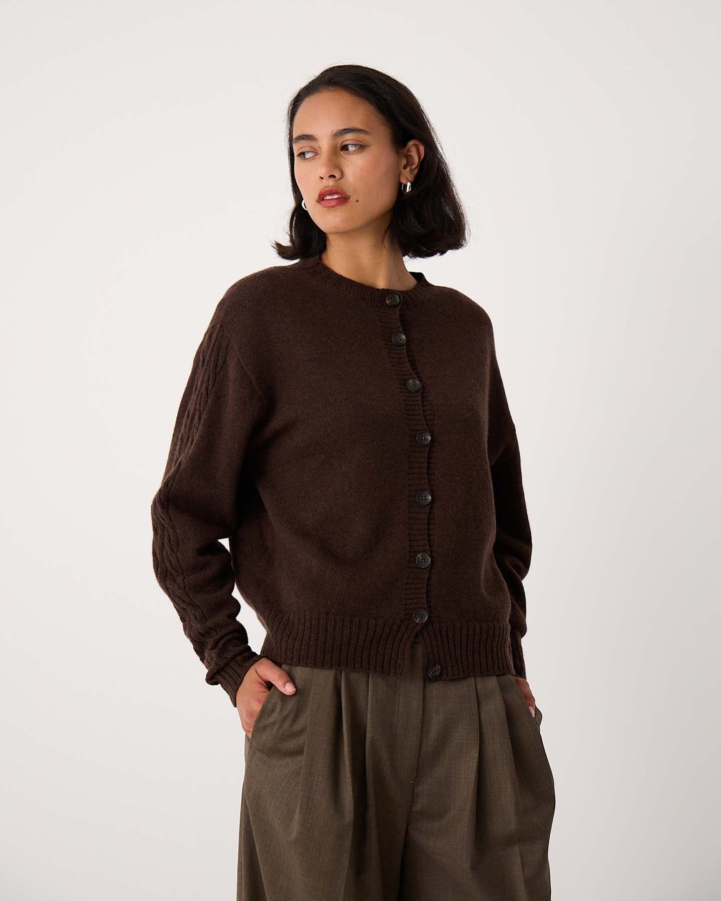 A woman wears the Kindred Cable Cardigan in Cocoa, styled with loose brown trousers, standing with one hand in her pocket against a plain white background.