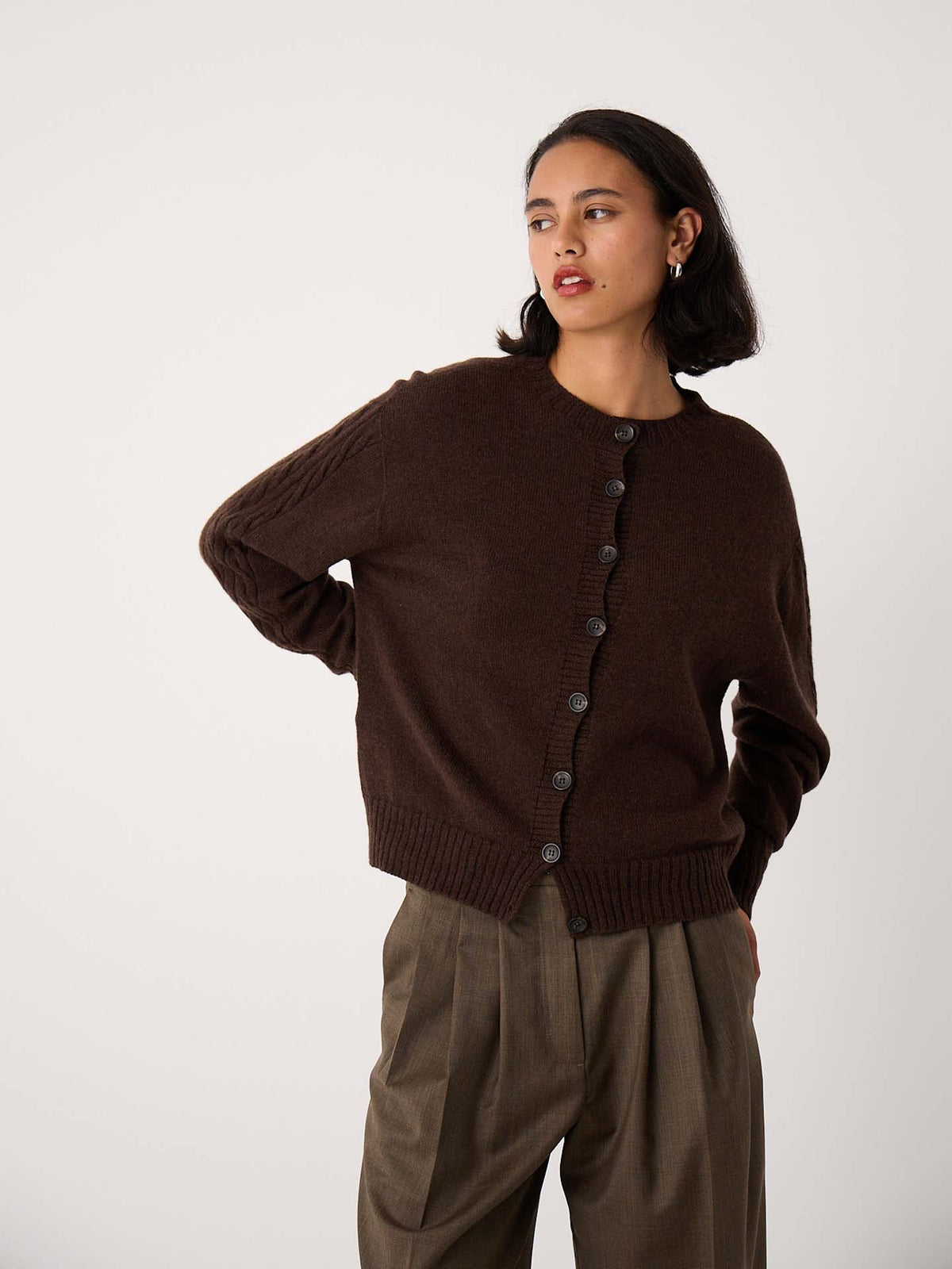A woman with short dark hair wears the Kindred Cable Cardigan – Cocoa and relaxed-fit brown trousers, standing against a plain light background.