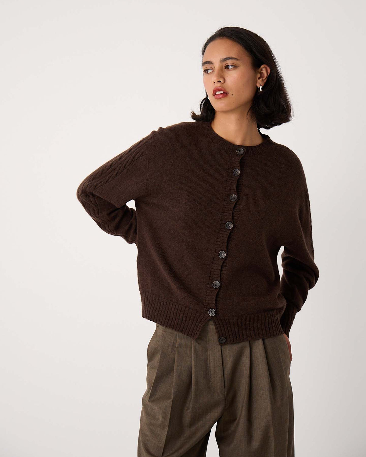 A woman with short dark hair wears the Kindred Cable Cardigan – Cocoa and relaxed-fit brown trousers, standing against a plain light background.