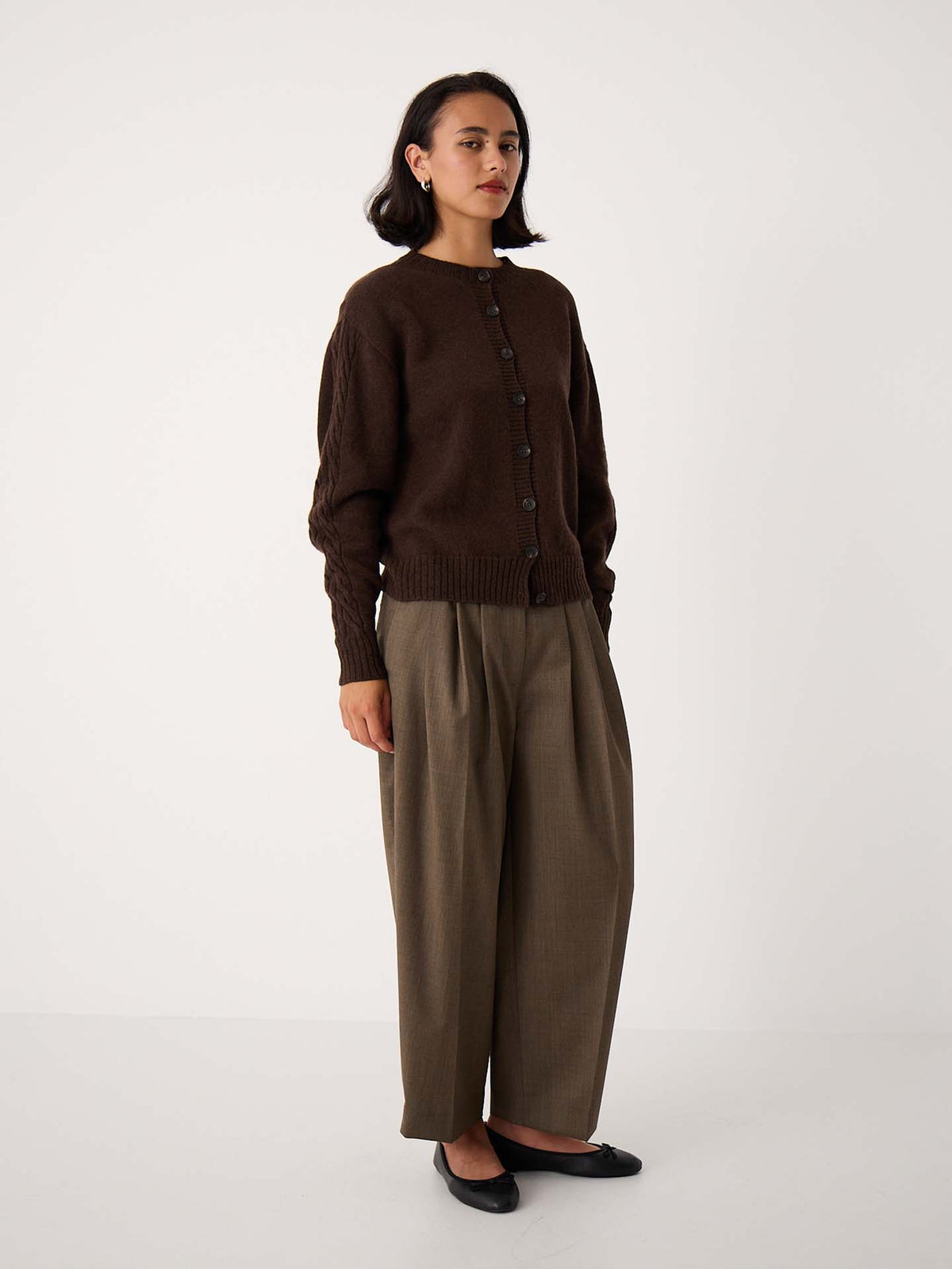 A woman stands against a plain background wearing the Kindred Cable Cardigan in Cocoa, relaxed taupe trousers, and black flats.