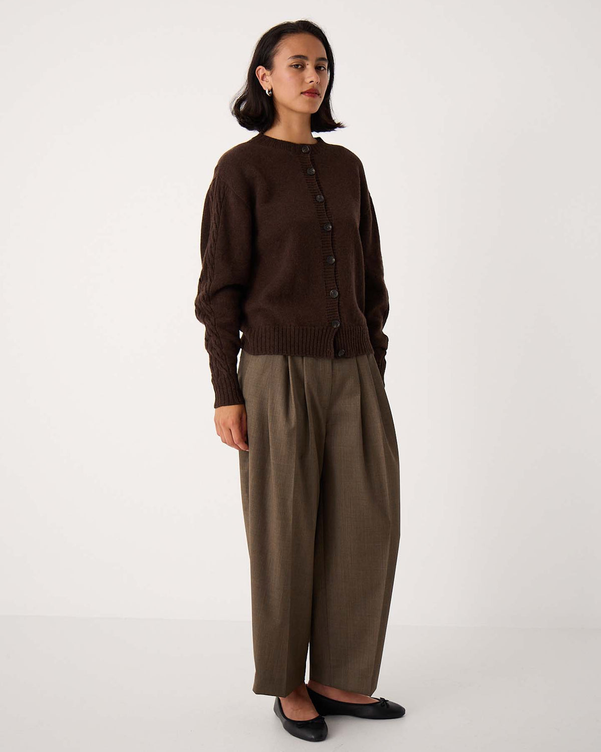 A woman stands against a plain background wearing the Kindred Cable Cardigan in Cocoa, relaxed taupe trousers, and black flats.