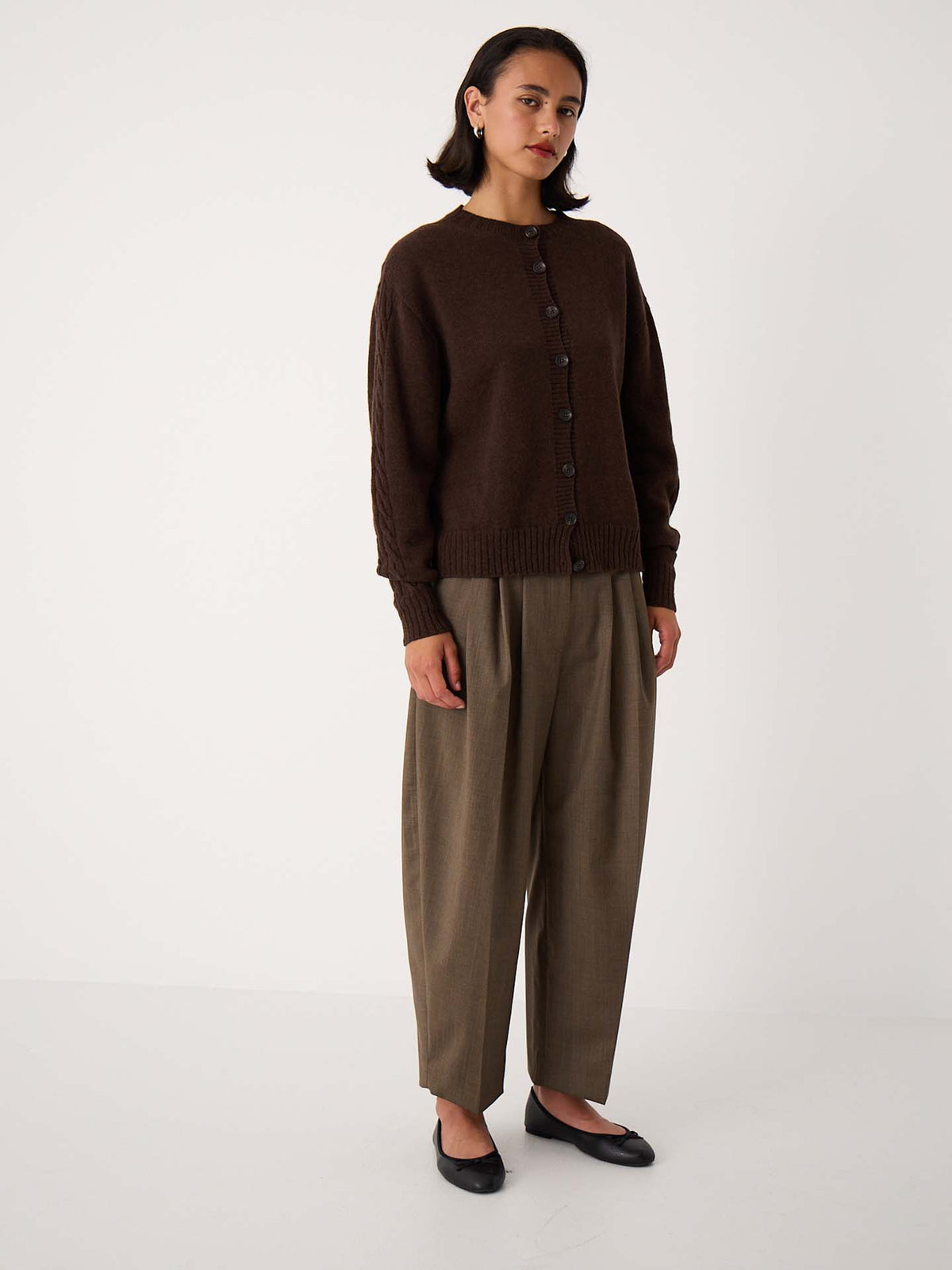 A woman stands against a plain white background wearing the Kindred Cable Cardigan – Cocoa, relaxed-fit olive-brown trousers, and black flats.