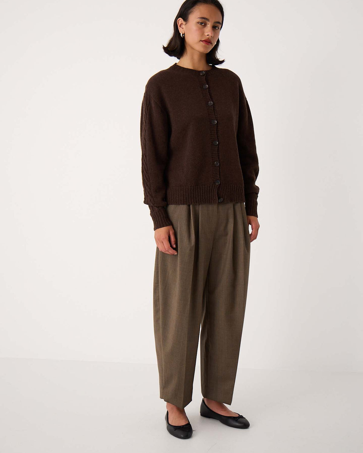 A woman stands against a plain white background wearing the Kindred Cable Cardigan – Cocoa, relaxed-fit olive-brown trousers, and black flats.