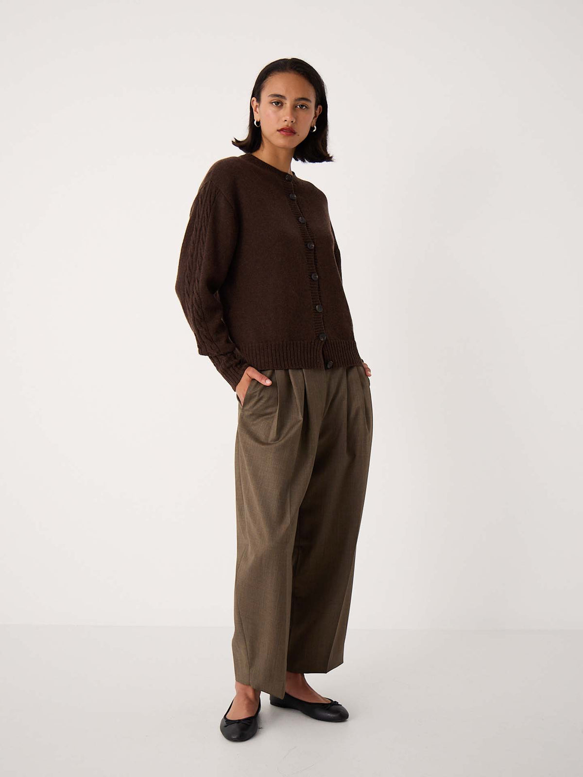 A person stands against a plain background wearing the Kindred Cable Cardigan – Cocoa in a relaxed fit, loose olive-green trousers, and black flats.