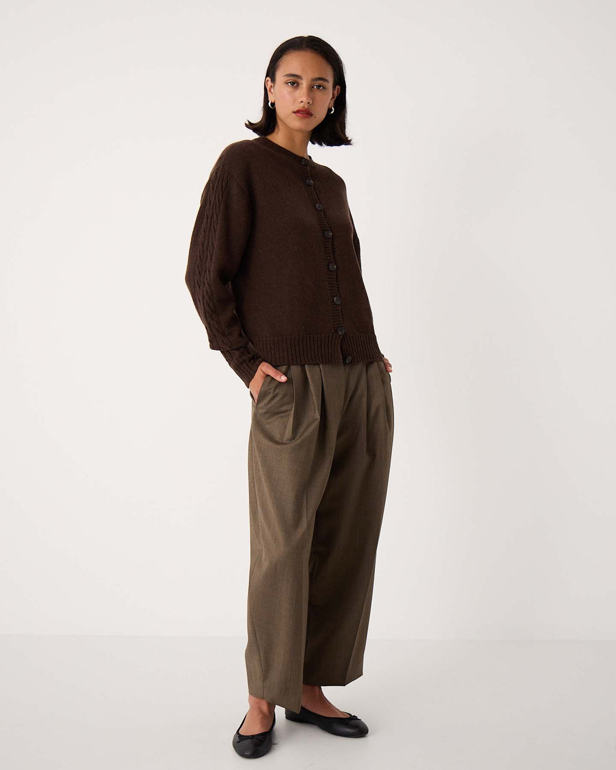 A person stands against a plain background wearing the Kindred Cable Cardigan – Cocoa in a relaxed fit, loose olive-green trousers, and black flats.
