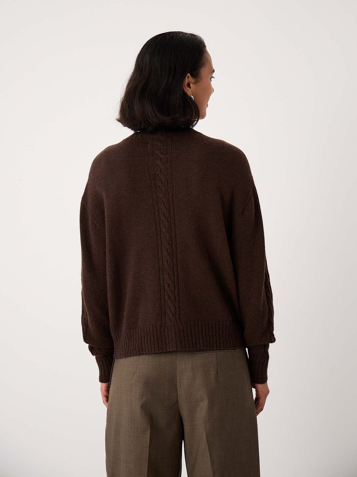 A person with short dark hair, seen from behind, wearing the Kindred Cable Cardigan – Cocoa and brown trousers against a plain light background.