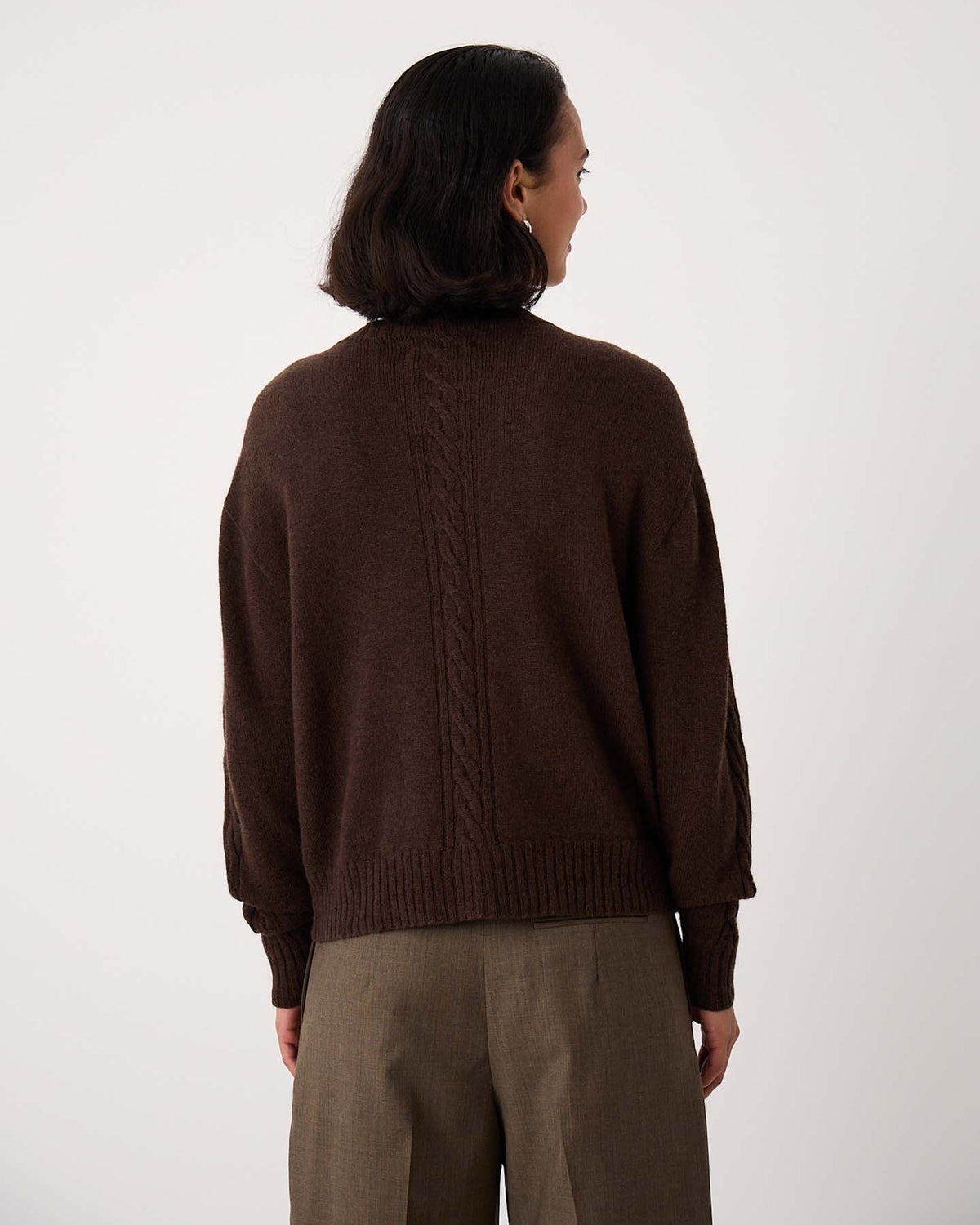 A person with short dark hair, seen from behind, wearing the Kindred Cable Cardigan – Cocoa and brown trousers against a plain light background.