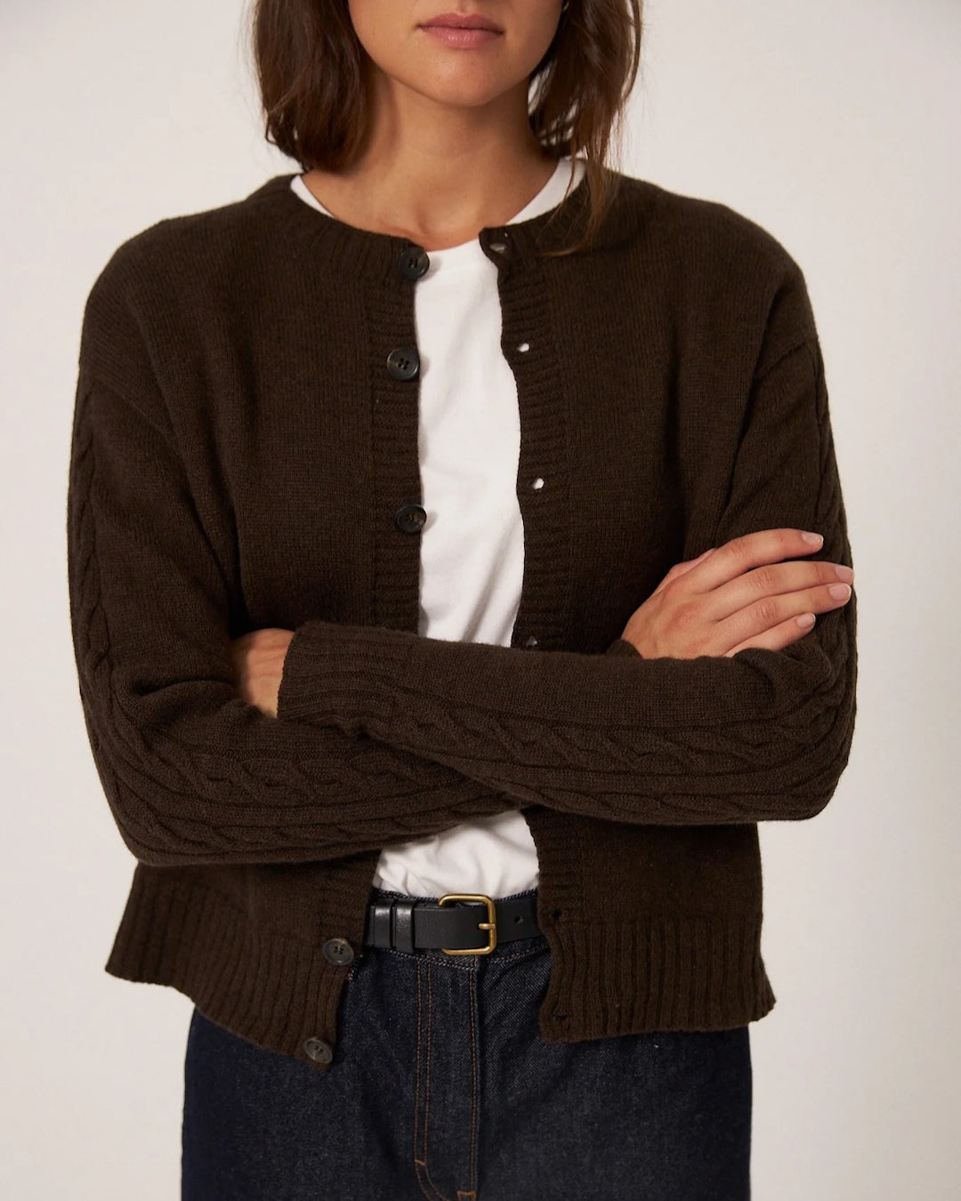 Dressed in Francie's Kindred Cable Cardigan in Cocoa over a white t-shirt, this individual exudes confidence with arms crossed. They complete the effortlessly stylish look with dark jeans and a black belt.
