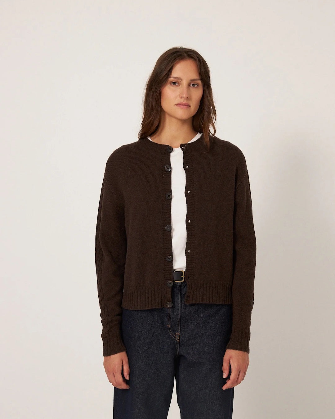A petite person models the Francie Kindred Cable Cardigan in Cocoa, a relaxed-fit dark brown piece, layered over a white shirt and paired with dark jeans, against a plain backdrop.