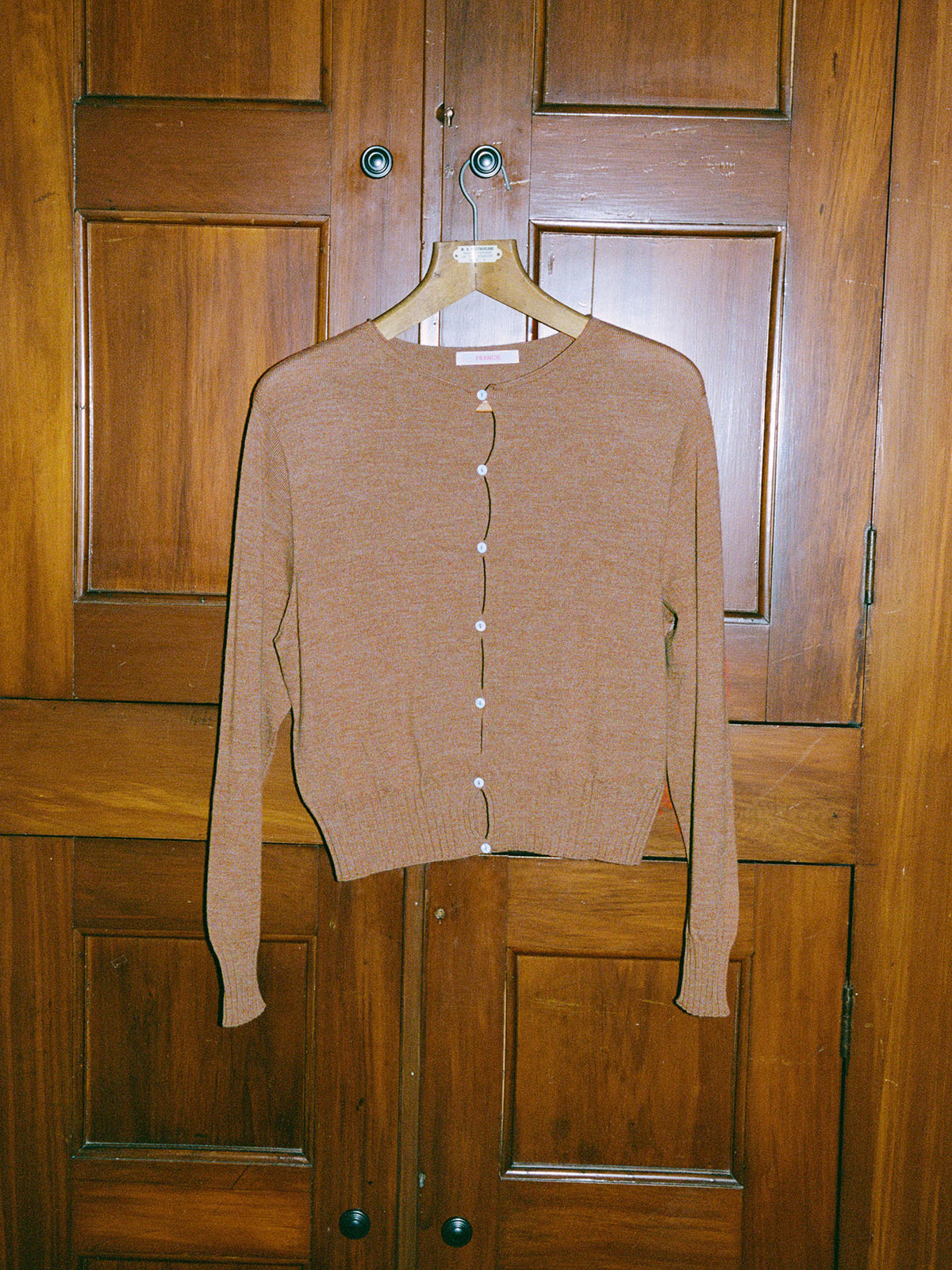 The Francie Anni Cardigan – Brown Sugar, a relaxed-fit long-sleeve cardigan with white buttons, hangs on a wooden hanger against a wood-paneled door.