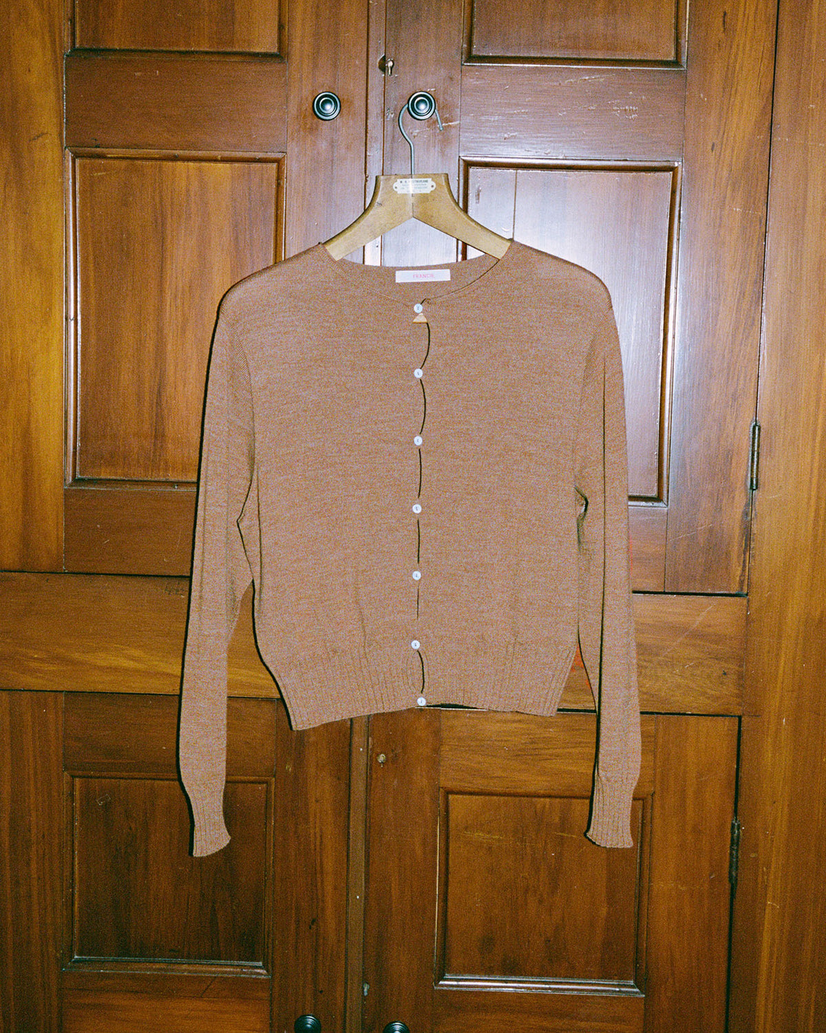 The Francie Anni Cardigan – Brown Sugar, a relaxed-fit long-sleeve cardigan with white buttons, hangs on a wooden hanger against a wood-paneled door.