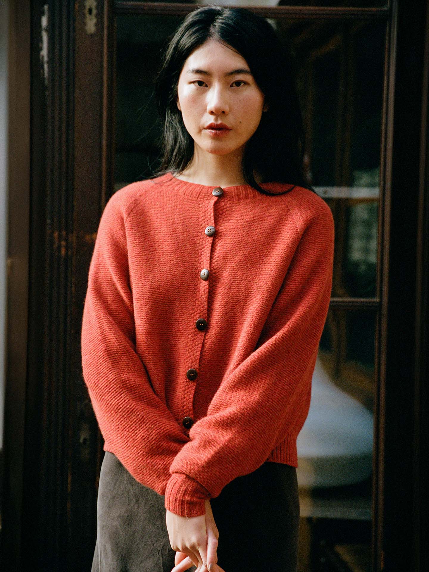 A woman with long dark hair stands indoors by a wooden door and window, wearing a brown skirt and the Loft Cardigan – Amber.