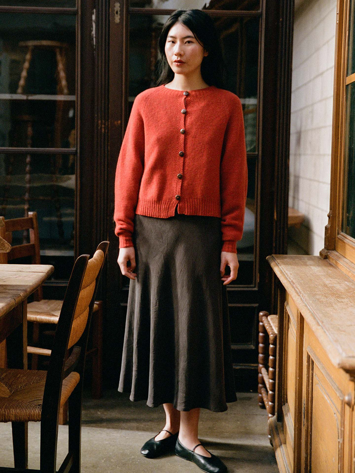 A woman stands indoors wearing a Loft Cardigan – Amber in a petite fit, styled with a long dark skirt and black shoes. She is in a room featuring wooden furniture and glass-fronted cabinets.