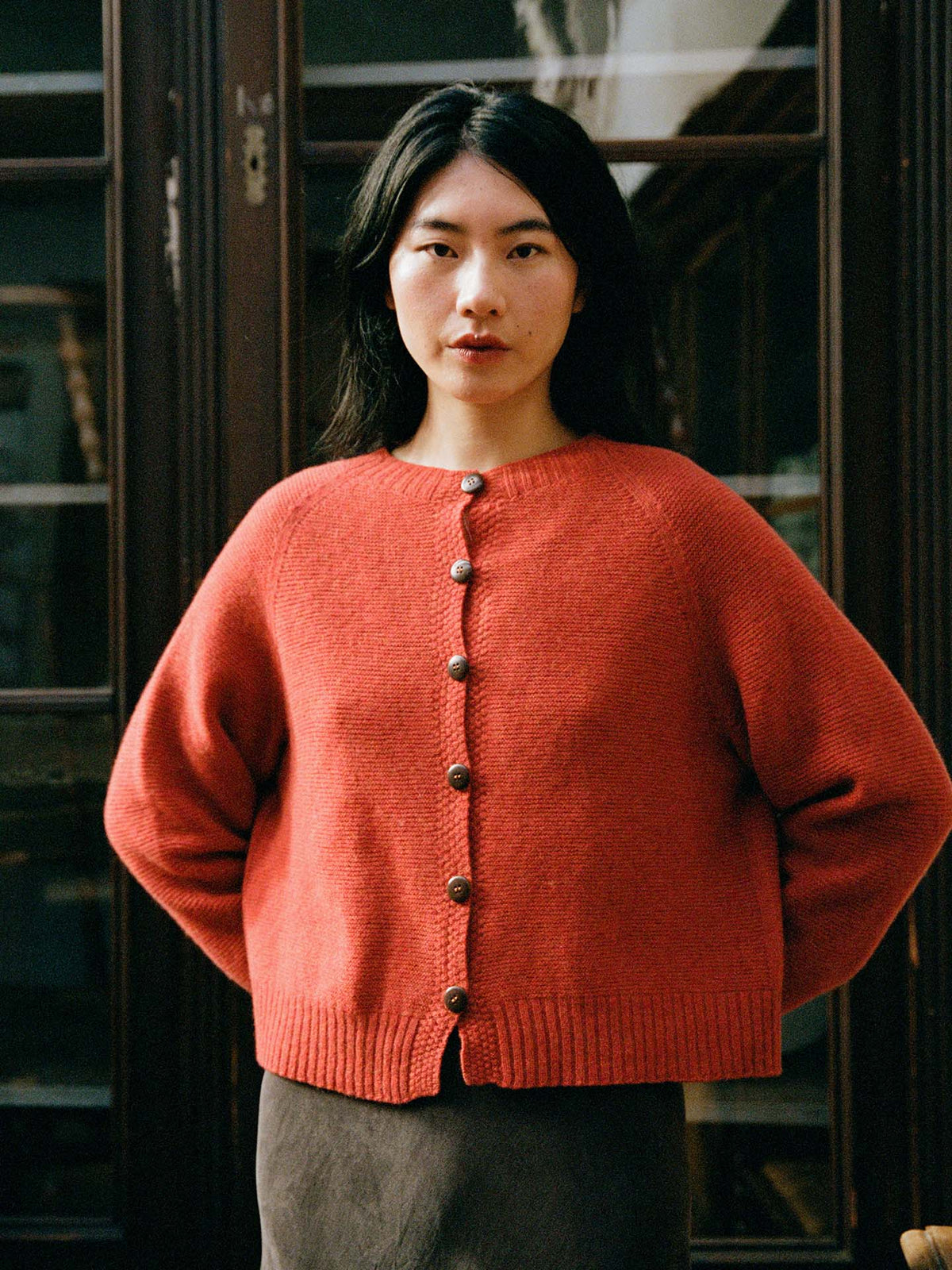 A woman stands indoors wearing a petite-fit Loft Cardigan – Amber and a dark skirt, hands behind her back, looking directly at the camera.