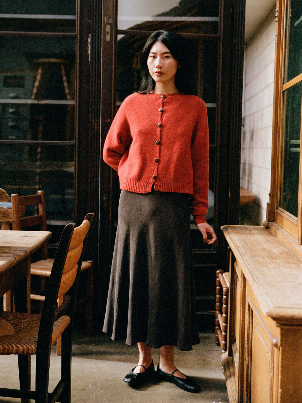 A woman stands indoors wearing a petite fit Loft Cardigan – Amber, a long brown skirt, and black shoes, surrounded by wooden chairs and cabinets.