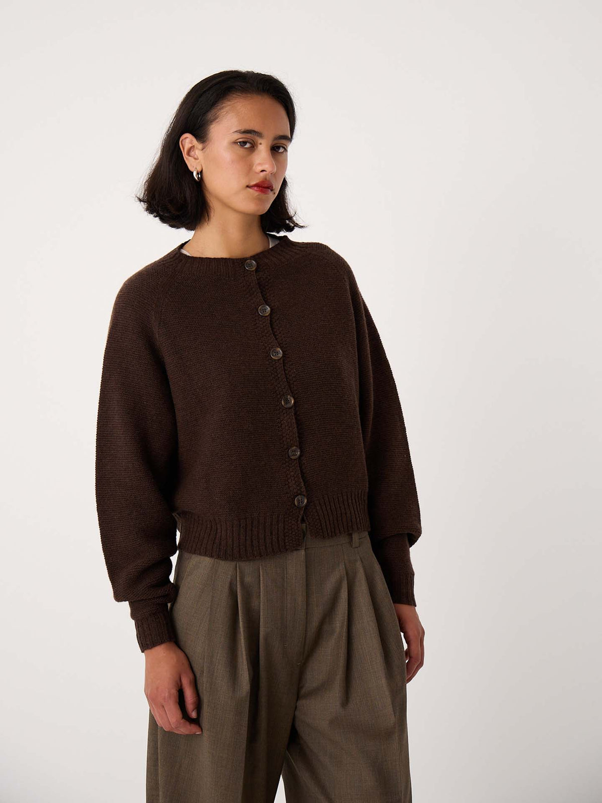 A woman with short dark hair wears the Loft Cardigan – Cocoa, styled buttoned over high-waisted loose khaki trousers, standing against a plain light background.