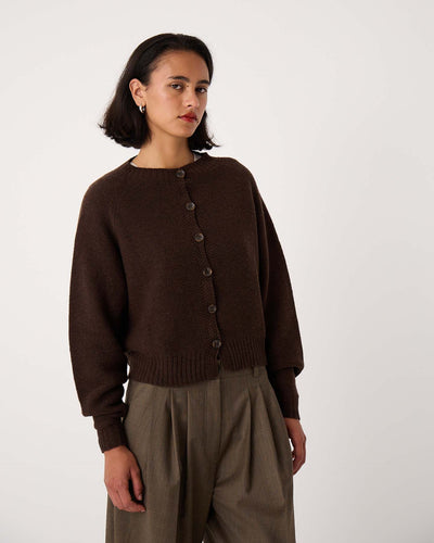 A woman with short dark hair wears the Loft Cardigan – Cocoa, styled buttoned over high-waisted loose khaki trousers, standing against a plain light background.