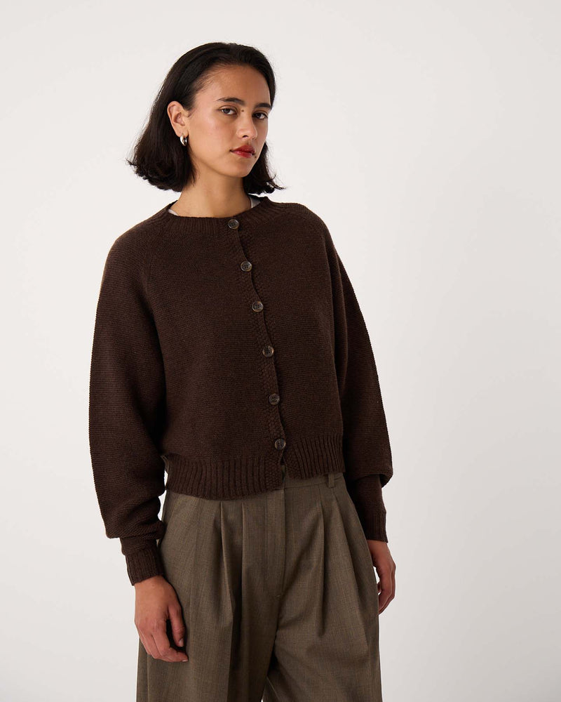 A woman with short dark hair wears the Loft Cardigan – Cocoa, styled buttoned over high-waisted loose khaki trousers, standing against a plain light background.