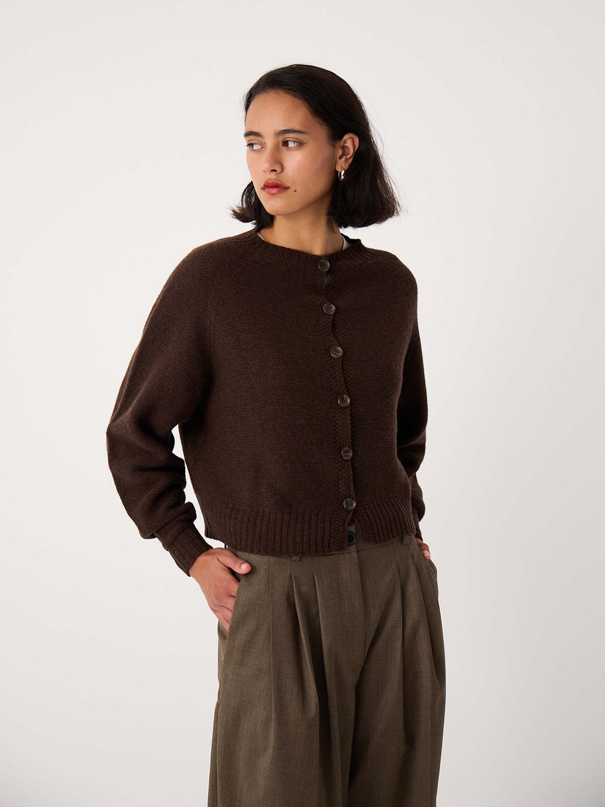 A woman in a Loft Cardigan – Cocoa and olive-green pants stands against a plain white background, looking to the side.