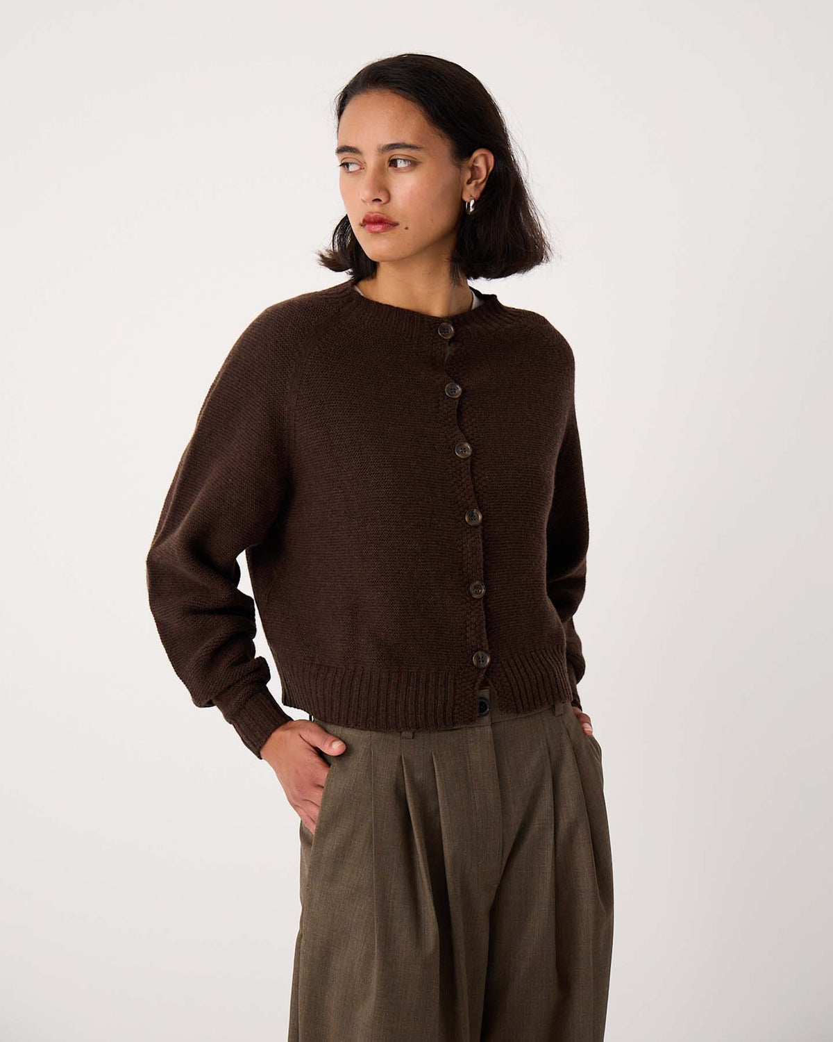 A woman in a Loft Cardigan – Cocoa and olive-green pants stands against a plain white background, looking to the side.
