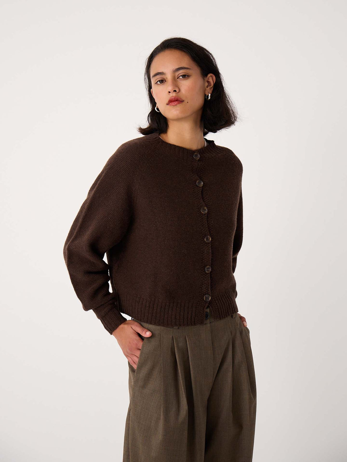 A woman stands against a plain background wearing the Loft Cardigan in Cocoa, paired with high-waisted, pleated khaki trousers.