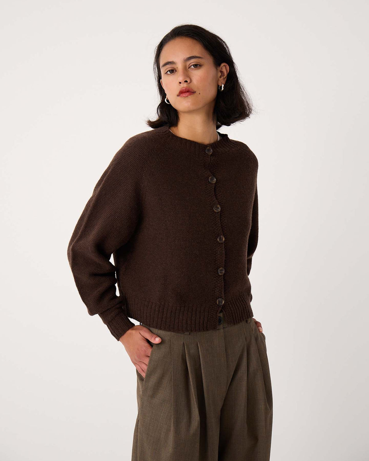 A woman stands against a plain background wearing the Loft Cardigan in Cocoa, paired with high-waisted, pleated khaki trousers.