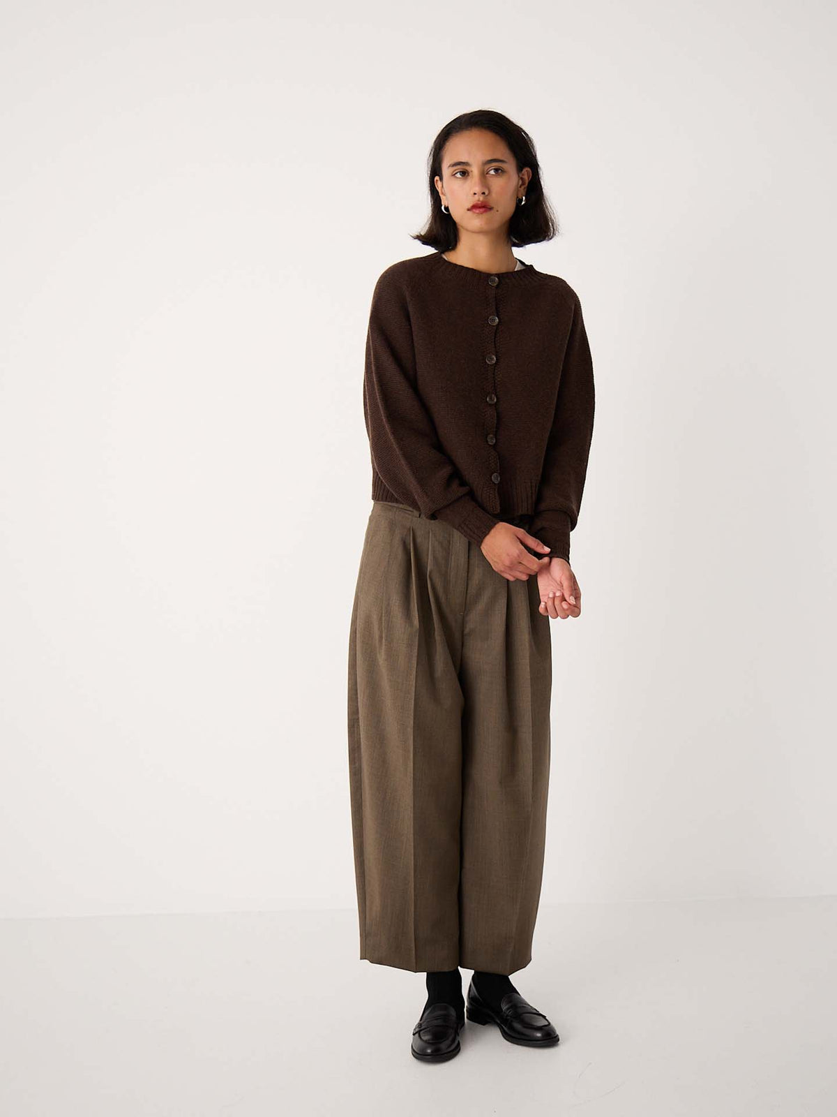A woman stands against a plain background wearing the Loft Cardigan – Cocoa, wide-leg olive trousers, and black shoes.