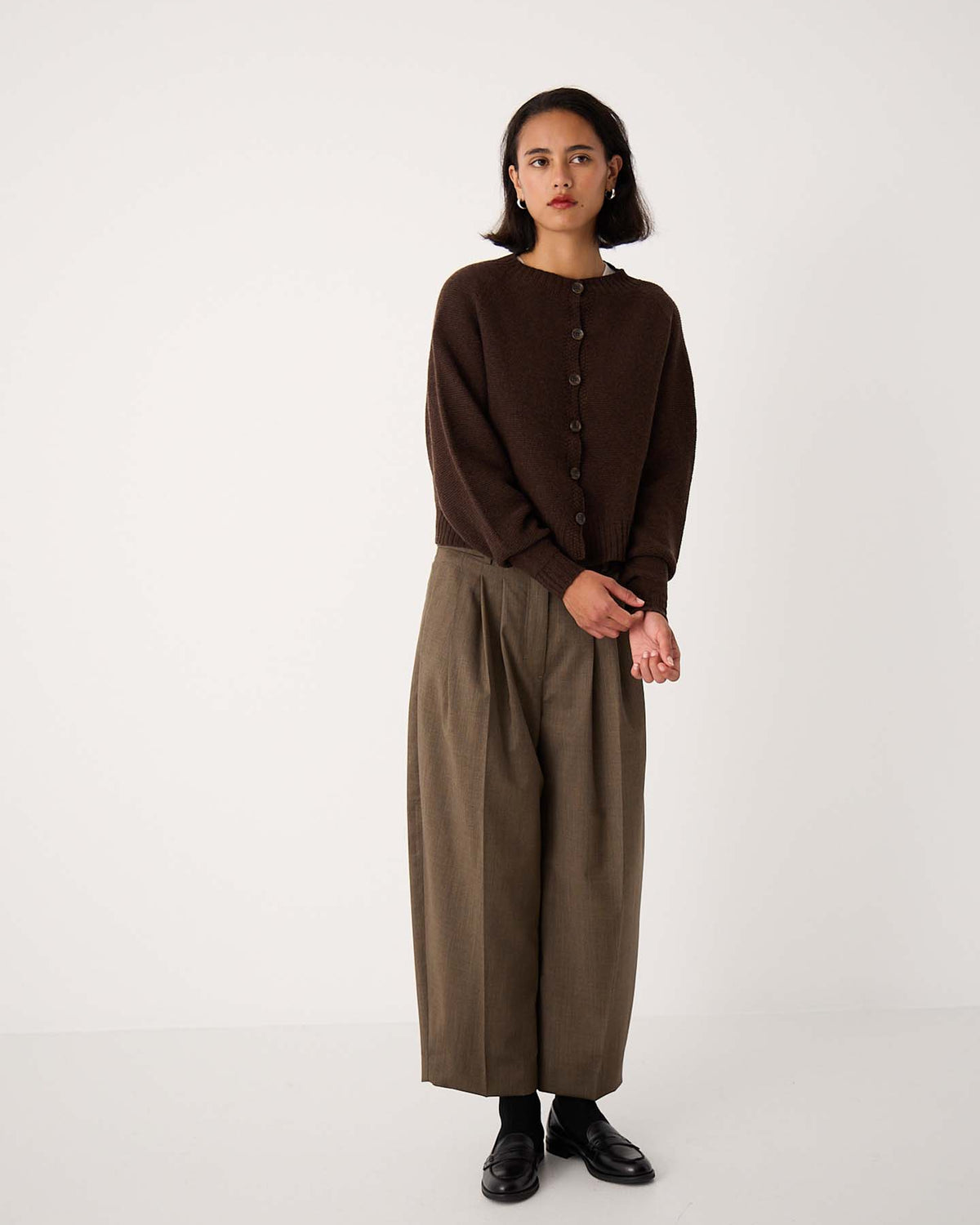 A woman stands against a plain background wearing the Loft Cardigan – Cocoa, wide-leg olive trousers, and black shoes.