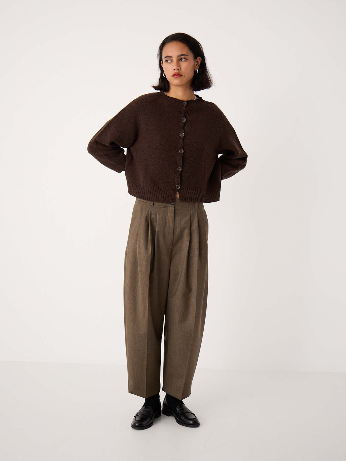 A person stands against a plain background wearing an oversized Loft Cardigan – Cocoa, loose brown pleated trousers, and black shoes.