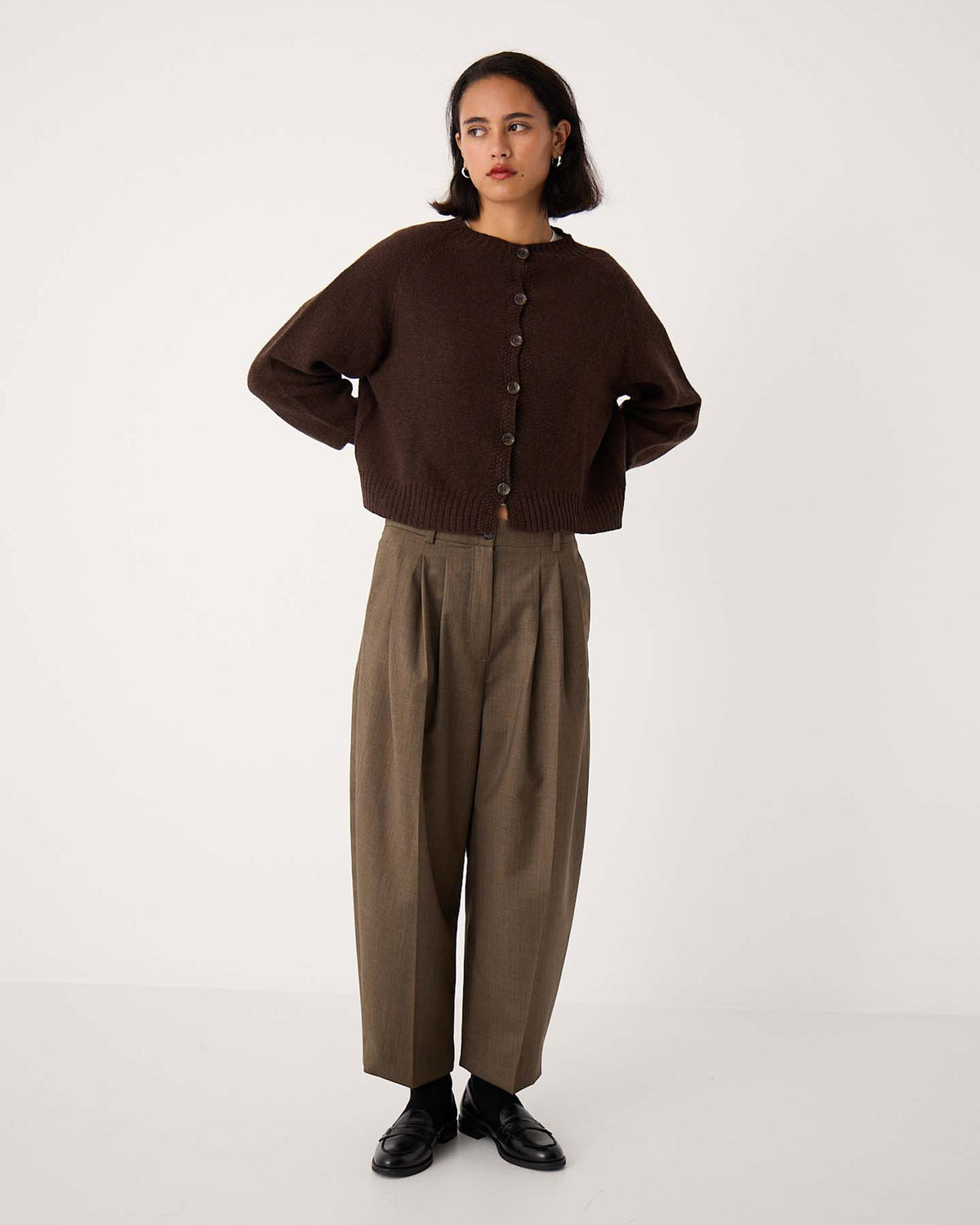 A person stands against a plain background wearing an oversized Loft Cardigan – Cocoa, loose brown pleated trousers, and black shoes.