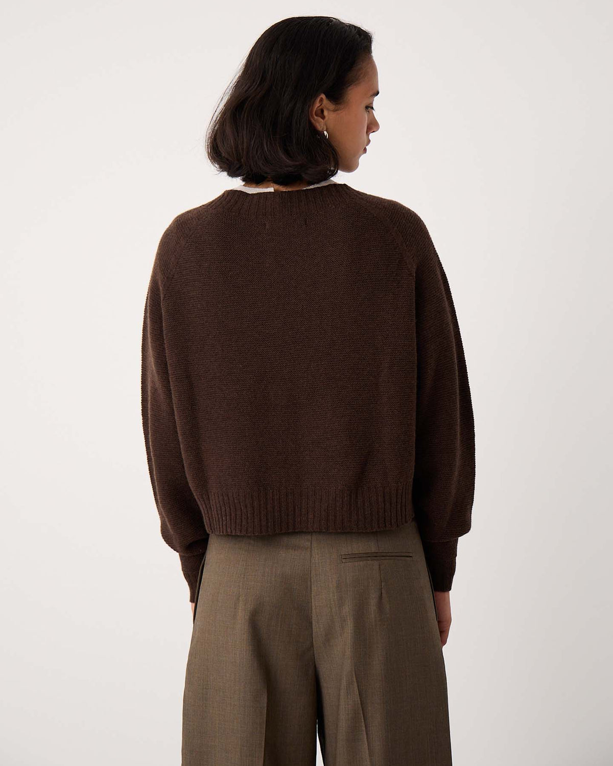 A person with short dark hair, wearing cocoa Loft Cardigan and khaki trousers, stands facing away from the camera against a plain background.