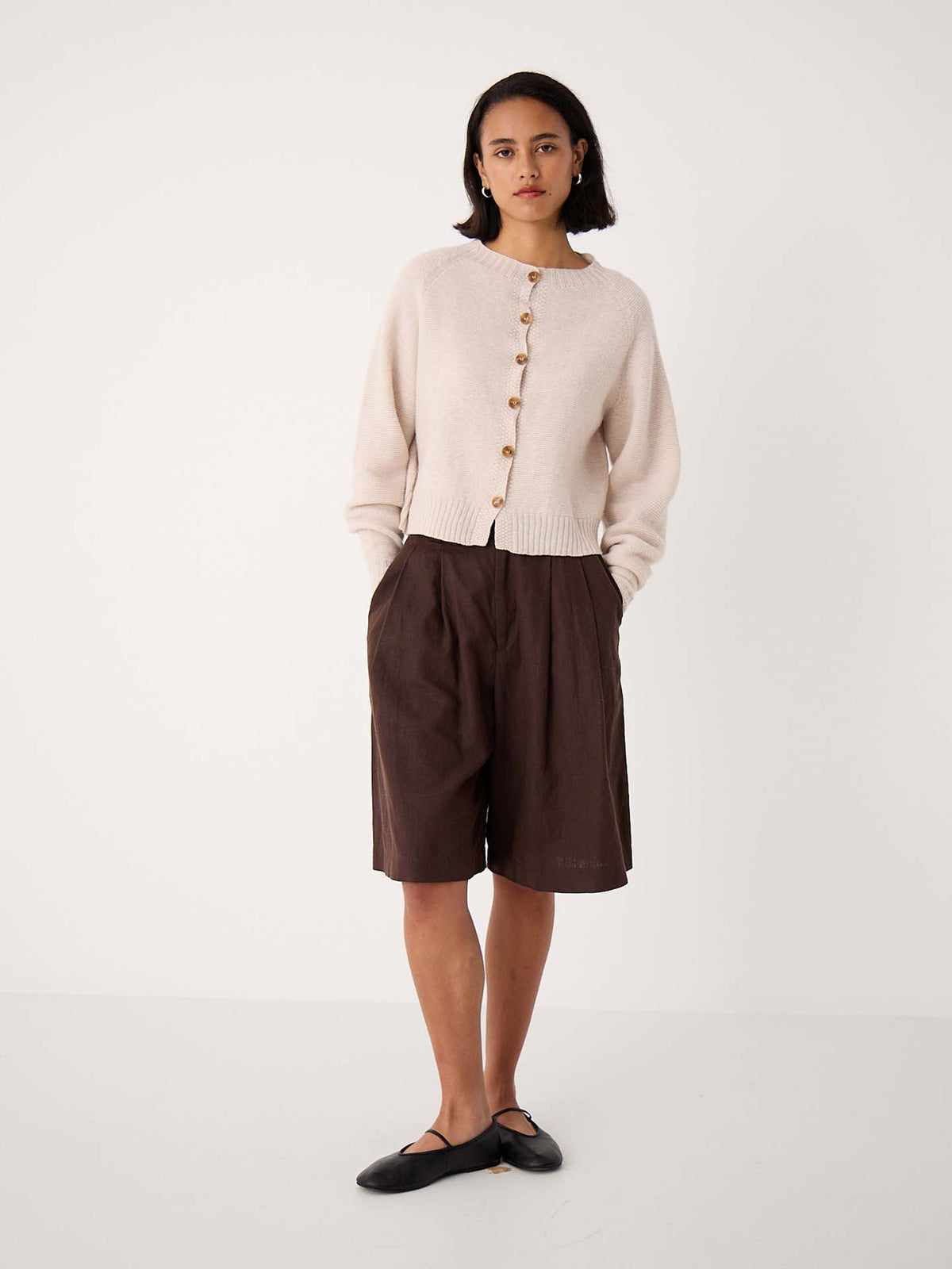 A person stands against a plain background wearing an oversized Loft Cardigan – Creme, dark brown knee-length shorts, and black flat shoes.