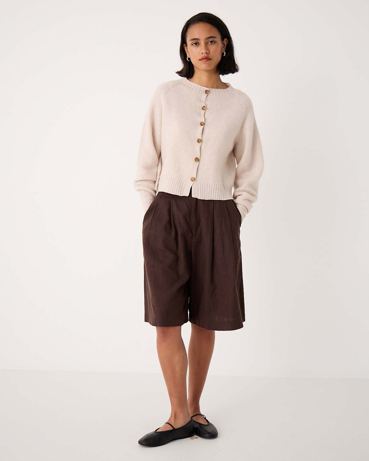 A person stands against a plain background wearing an oversized Loft Cardigan – Creme, dark brown knee-length shorts, and black flat shoes.