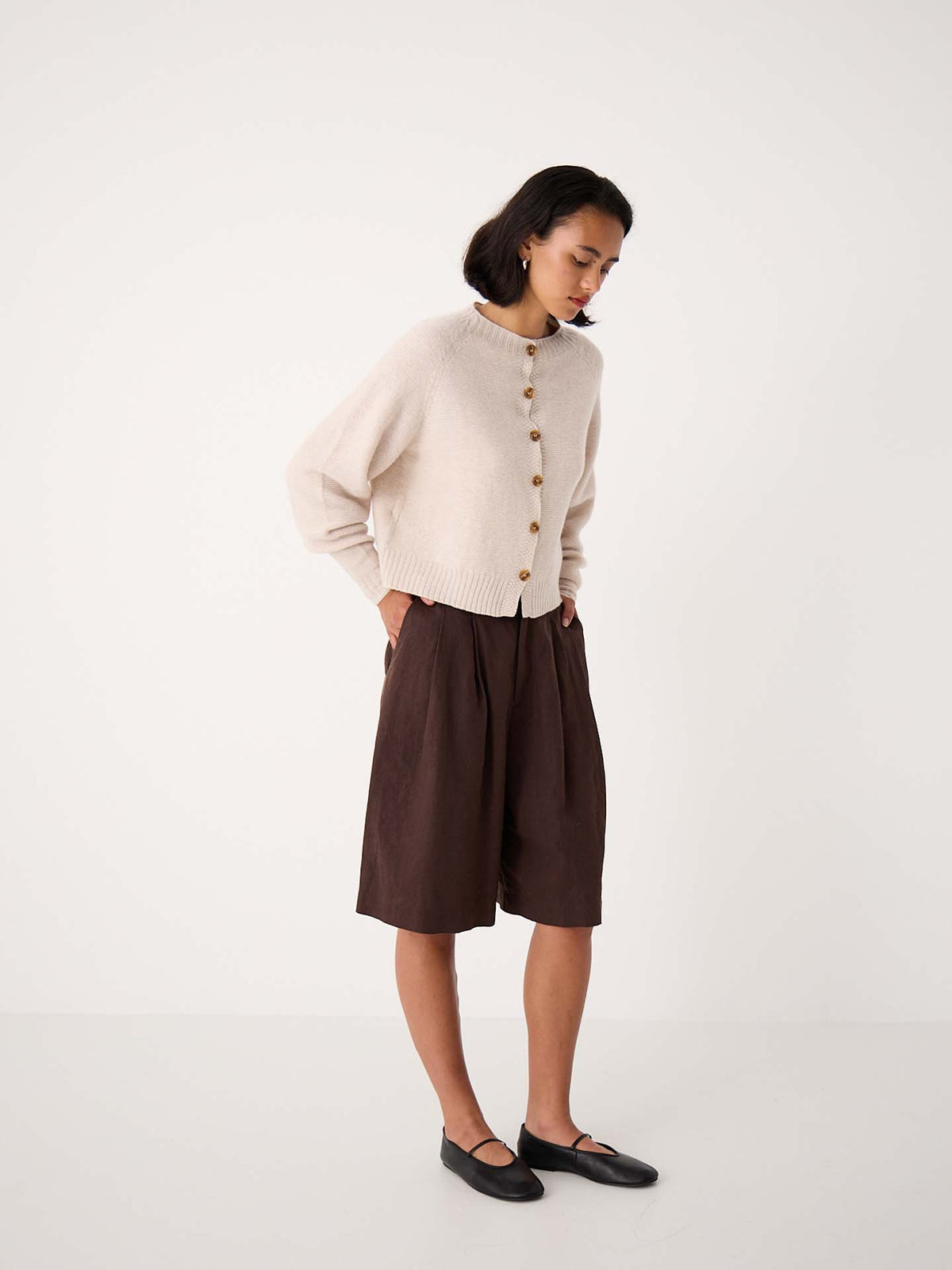 A woman stands indoors wearing a creme Loft Cardigan, loose brown knee-length shorts, and black flats, looking down with her hands in her pockets.