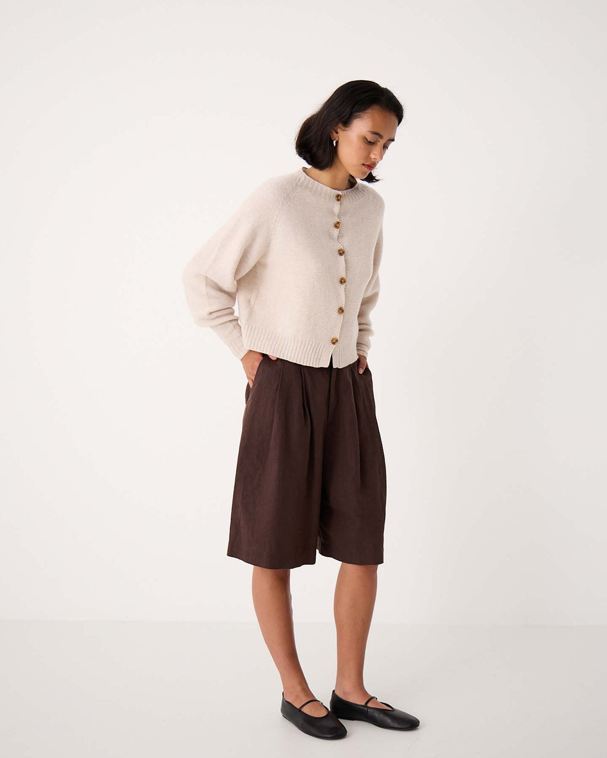 A woman stands indoors wearing a creme Loft Cardigan, loose brown knee-length shorts, and black flats, looking down with her hands in her pockets.
