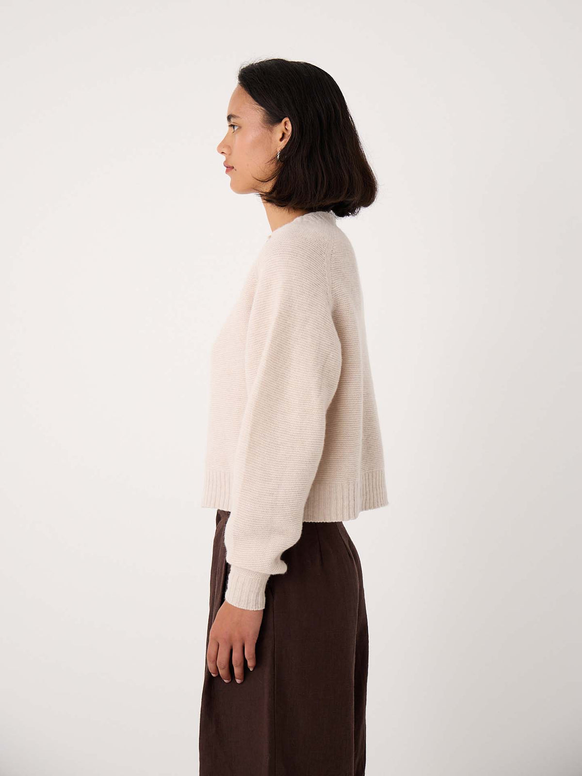 Woman standing in profile against a plain background, wearing the Loft Cardigan – Creme and a dark brown skirt.