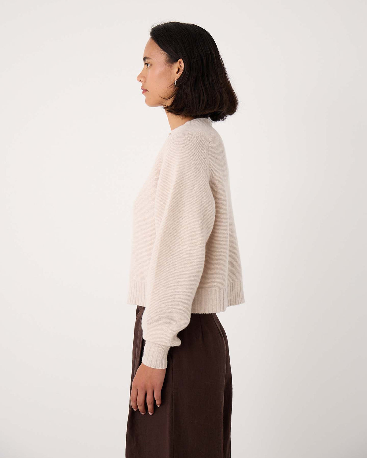 Woman standing in profile against a plain background, wearing the Loft Cardigan – Creme and a dark brown skirt.
