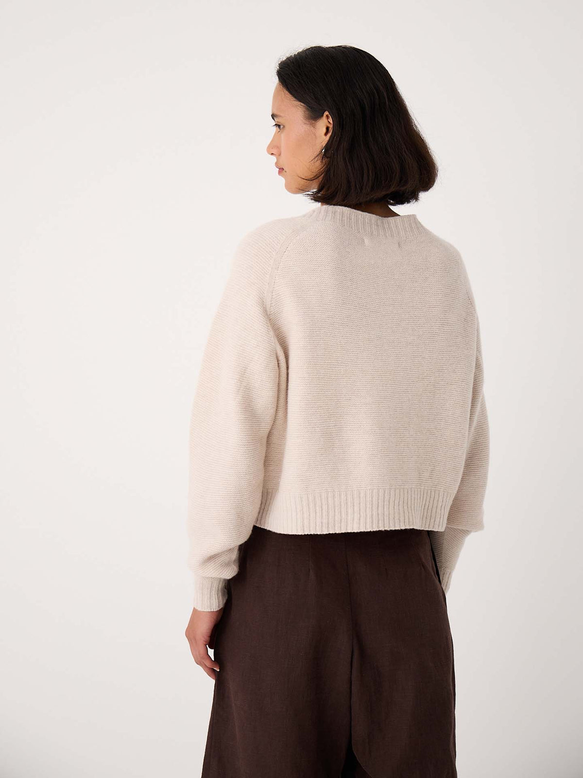 A woman with short dark hair, wearing an oversized Loft Cardigan – Creme and dark brown pants, stands facing away from the camera against a plain light background.