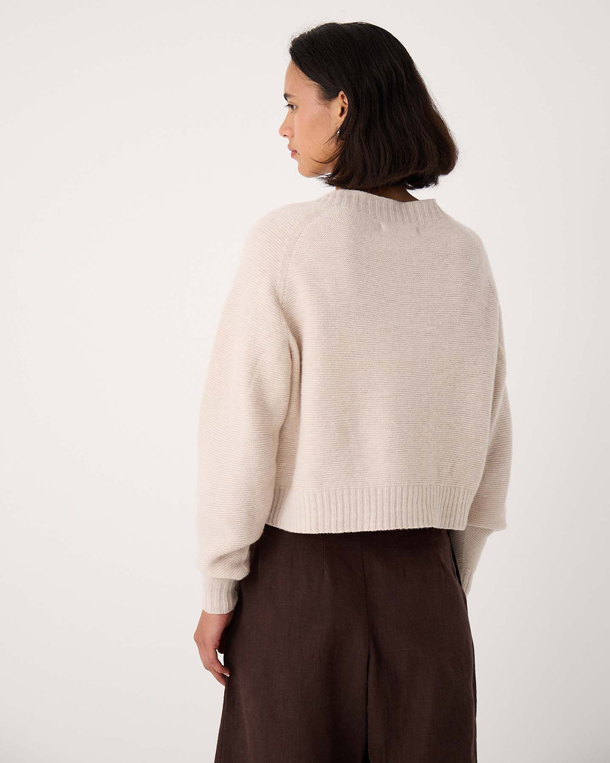 A woman with short dark hair, wearing an oversized Loft Cardigan – Creme and dark brown pants, stands facing away from the camera against a plain light background.
