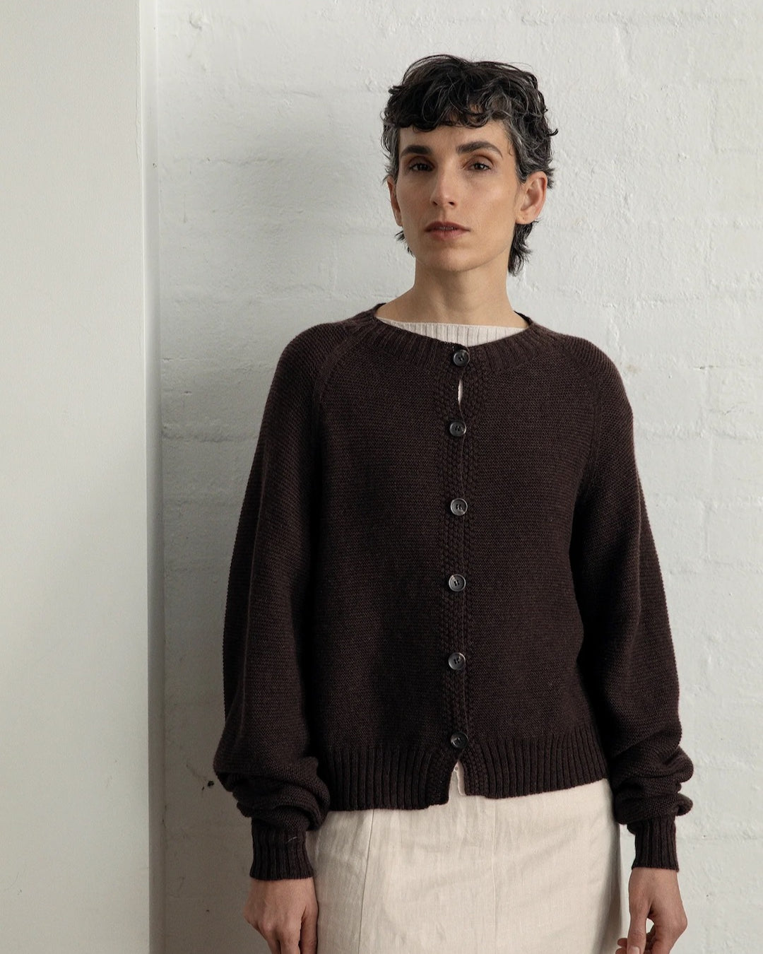 A person with short dark hair stands against a white wall, wearing a Francie Loft Cardigan in Cocoa and a light-colored skirt.