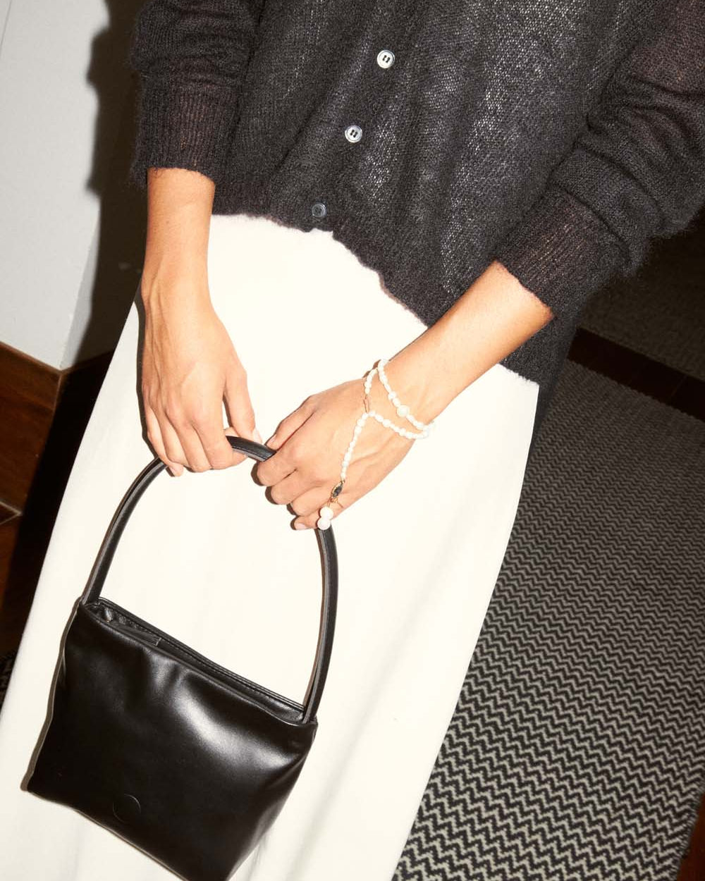 Wearing a Mist Cardigan – Black in a relaxed fit and a white skirt, a person holds a small black handbag with a beaded wrist strap, standing on a patterned carpet.