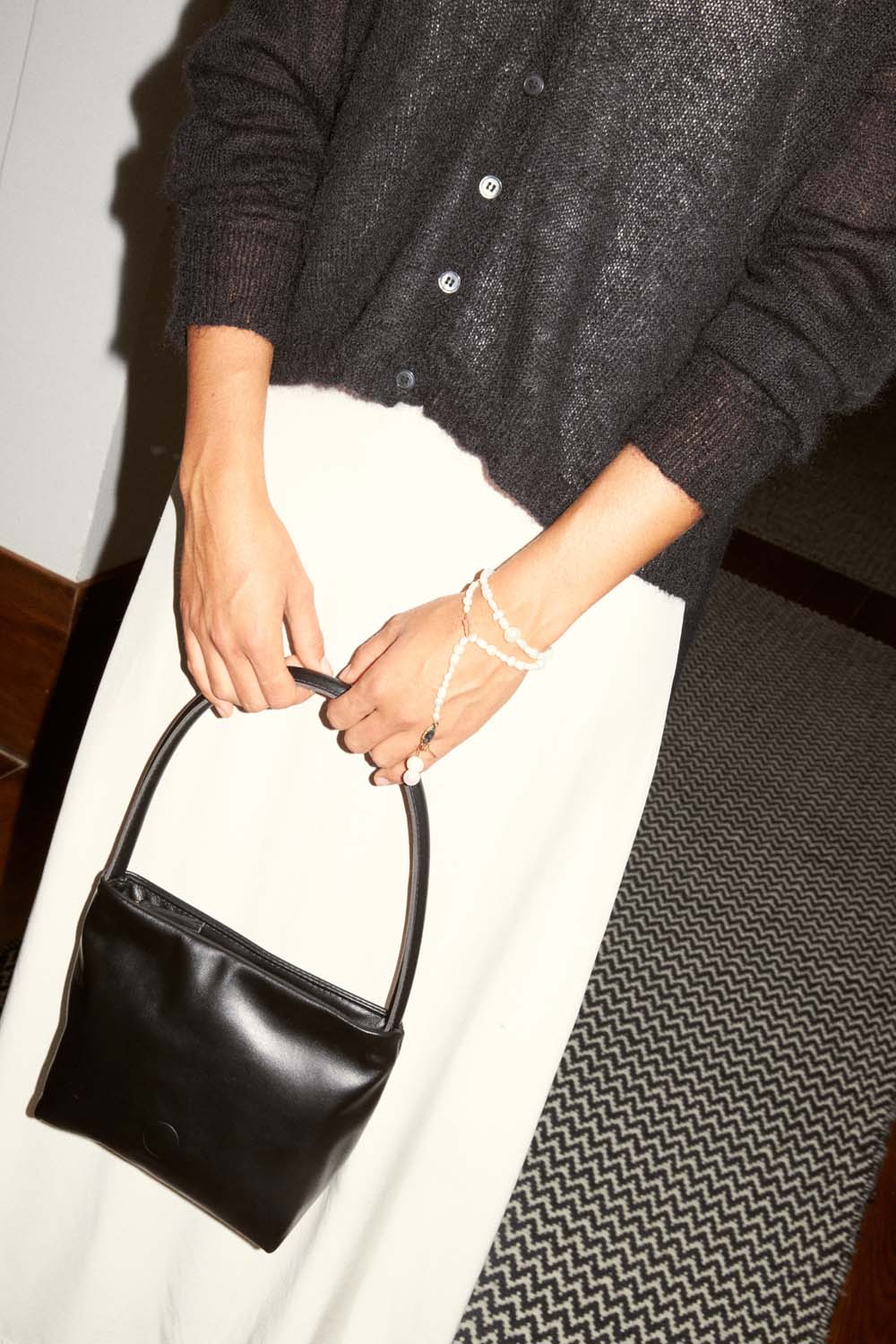 Wearing a Mist Cardigan – Black in a relaxed fit and a white skirt, a person holds a small black handbag with a beaded wrist strap, standing on a patterned carpet.