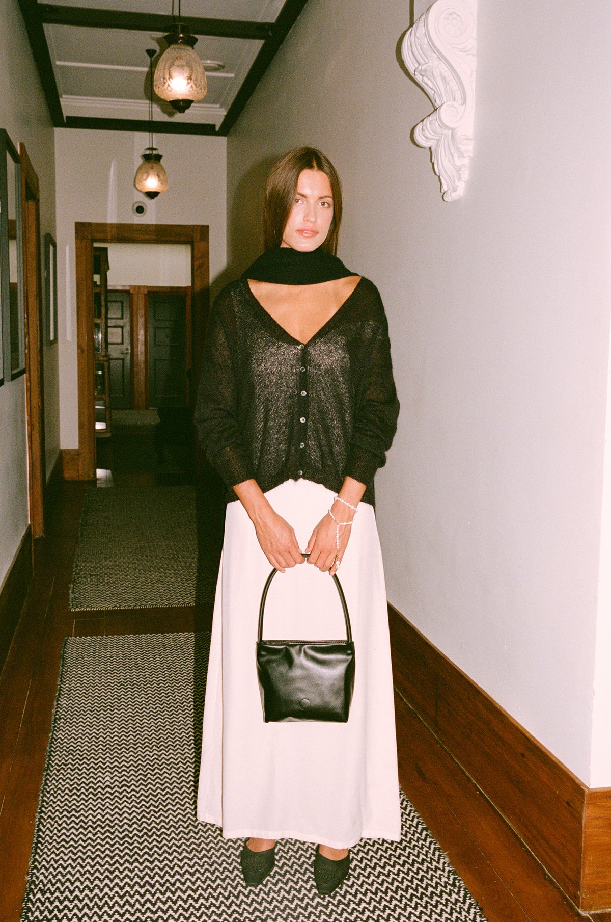 A petite woman stands in a hallway wearing the Mist Cardigan – Black, a relaxed-fit black top, paired with a white skirt, black scarf, black shoes, and holding a black handbag. The hallway has patterned rugs, white walls, and hanging lights.