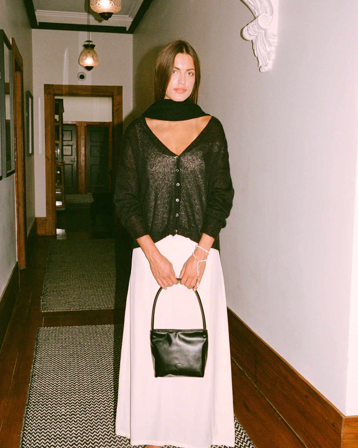 A petite woman stands in a hallway wearing the Mist Cardigan – Black, a relaxed-fit black top, paired with a white skirt, black scarf, black shoes, and holding a black handbag. The hallway has patterned rugs, white walls, and hanging lights.